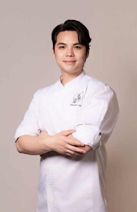 Chef Vincent Lai mặc áo bếp trắng, khoanh tay và mỉm cười trước nền màu be · Chef Vincent Lai in a white chef's jacket, arms folded and smiling, against a beige background
