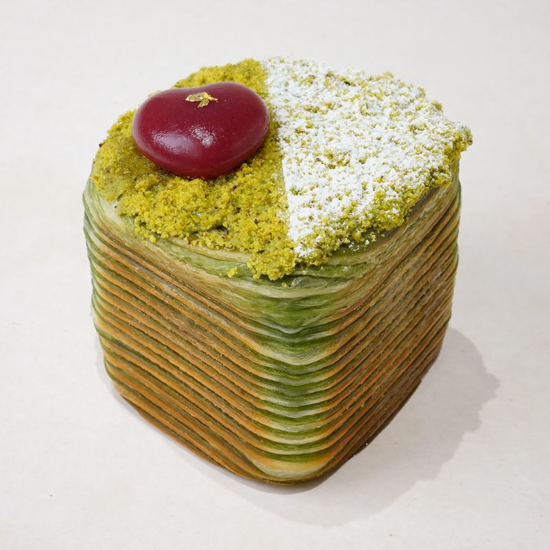 Emerald Pistachio · Bánh ngàn lớp màu xanh lá, thành bánh lộ rõ các lớp bột, mặt phủ vụn xanh, đường bột và một viên tròn màu đỏ sẫm bóng · Green laminated pastry with visible layers on its sides, topped with green crumb, icing sugar and a glossy dark red dome